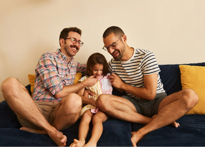 Two fathers sitting on a couch with their daughter in between them, all smiling and laughing.