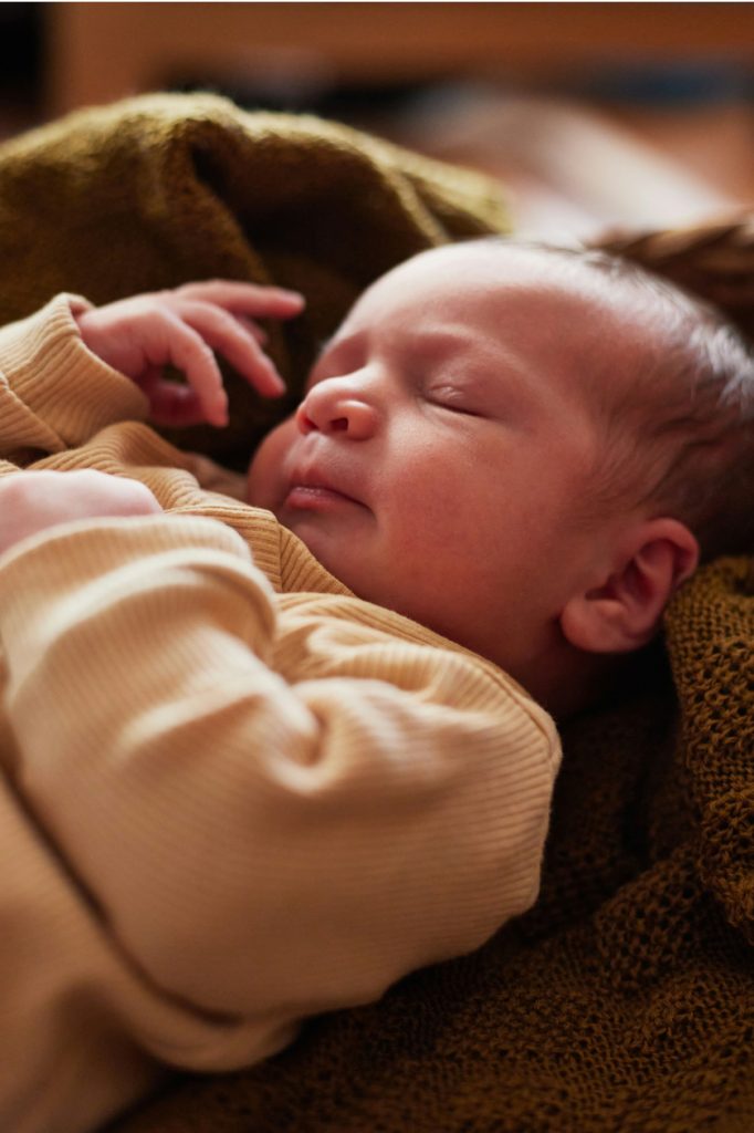 Newborn baby wearing a tan sweater sleeping on a blanket, looking peaceful.