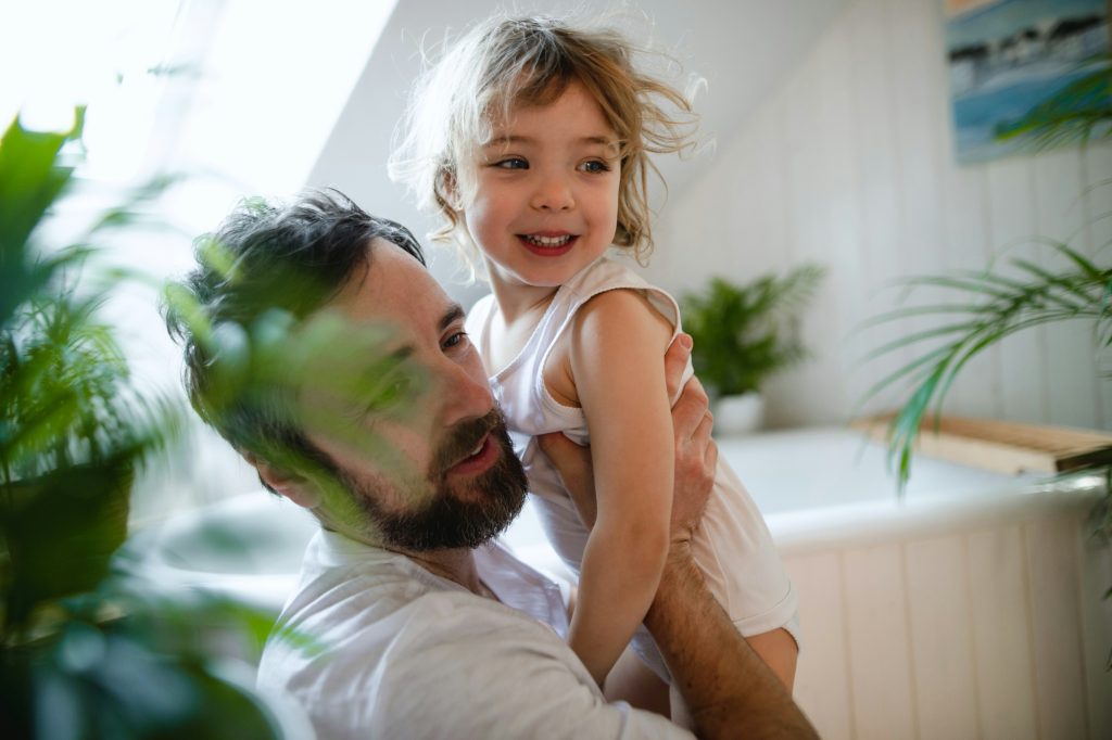 A young father holding his smiling daughter while both look at something across the room.